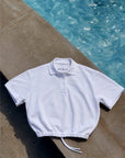 A white short-sleeve polo shirt with a drawstring hem, laid flat on a concrete poolside surface with blue water in the background.