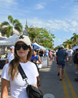 Woman wearing a white cap with a black logo and sunglasses, standing on a street with palm trees and people in the background.