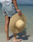 Person holding a straw hat on a beach with clear water and blue sky.