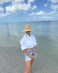 Person wearing a white shirt, denim shorts, and a straw hat standing on a beach with clear blue water and sky.