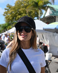 Woman wearing sunglasses and a black cap in an outdoor setting with tents and people in the background.
