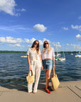 Two women standing on a concrete pier by a lake with boats and a blue sky in the background. The woman on the left wears a white lace jumpsuit and holds a beige tote bag.