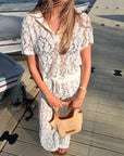 A woman wearing a white lace short-sleeve top and matching white lace maxi skirt stands on a wooden dock holding a woven straw handbag.