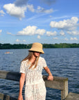 A woman wearing a straw bucket hat and a white lace short-sleeve dress stands on a wooden dock by a lake with a tree-lined shore and blue sky with clouds in the background.