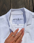 A close-up of a white polo shirt laid flat on a dark wooden surface, with a hand touching the collar area. The inside neck label reads ANYA & NIKI.