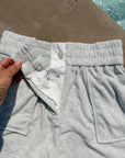 Close-up of light gray heather sweatpants with a hand holding the waistband, showing a white inner lining and snap-button closure, with a poolside concrete surface in the background.