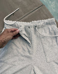A close-up of light gray heather sweatpants with a hand adjusting the drawstring waist. The shorts are shown outdoors next to a concrete surface and water, featuring a front zipper, drawstrings, and a side pocket.