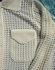 Close-up of a cream-colored open-knit cardigan sweater with a small chest pocket, showing the textured knit pattern and collar.