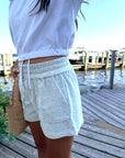 A person standing on a wooden dock by the water wearing light heather gray drawstring shorts and a white cropped top, holding a woven straw bag.