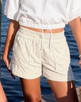 A person wearing light beige textured drawstring shorts and a white cropped top, standing outdoors with water in the background.
