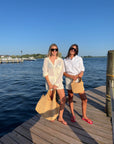 Two women standing on a wooden dock by the water, wearing sunglasses, white tops, and straw tote bags. The background features a clear blue sky and calm water.