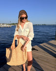 A woman stands on a wooden dock by the water wearing a cream-colored crochet knit shirt and shorts set, black sunglasses, and carrying a large woven straw tote bag.