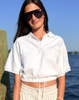 A woman wearing a white short-sleeve cropped polo shirt and light beige drawstring sweatpants, standing outdoors near the water with a wooden post on the left and blue sky and water in the background.