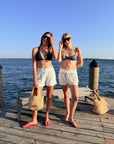 Two women posing on a wooden dock by the water, wearing matching bikini tops and white shorts. One wears a black bikini top and light gray shorts, while the other wears a gray bikini top and cream shorts.