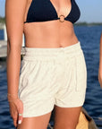 A close-up of a person outdoors by the water wearing light heather grey shorts and a dark navy bikini top with a small gold ring detail at the center.