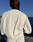 A person viewed from behind wearing a light heather gray sweatshirt with a small rectangular brand patch on the upper back, standing outdoors near water under a blue sky.