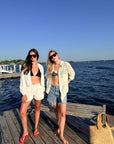 Two women posing on a wooden dock next to a body of water under a clear blue sky. One is wearing a white open-front jacket and grey shorts over a black bikini top, and the other is wearing a white open-front shirt and denim shorts over a gray bikini top.