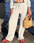 A person standing on a wooden dock by the water wearing light heather gray wide-leg drawstring sweatpants, a white cropped long-sleeve top, and red flip-flops, while holding a woven straw tote bag.