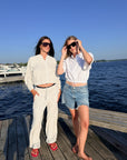 Two women standing on a wooden dock by the water wearing white summer outfits, with one in a white cropped zip-up hoodie and pants and the other in a white cropped button-up shirt with denim shorts. Both are wearing sunglasses.