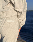 Close-up of a person wearing a light heather gray sweatshirt and matching jogger pants, with a small visible 'ANYA & NIKI' label on the pocket area. The background shows an outdoor water scene with blue sky and blurred steps.