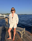 A woman wearing a light grey terry lounge set consisting of a long-sleeve button-up jacket and matching shorts, standing on a wooden dock with a lake and blue sky in the background.