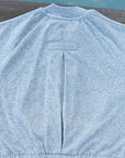 A light heather gray sweatshirt laid flat on a concrete surface with a swimming pool in the background. The back of the sweatshirt features a central seam and a small rectangular brand label near the collar.
