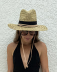 A woman wearing a woven straw fedora-style sun hat with a black band and beige chin cord, paired with dark sunglasses and a black halter top, standing against a textured white wall.