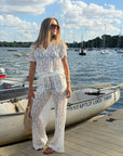 A woman stands on a wooden dock by a lake wearing a white lace short-sleeve jumpsuit with a drawstring waist and wide-leg pants. She is holding a straw bag, with a white boat and water in the background.