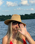 A woman with blonde hair wearing a straw sun hat, dark sunglasses, and a red top poses in front of a lake with trees and a blue sky in the background.
