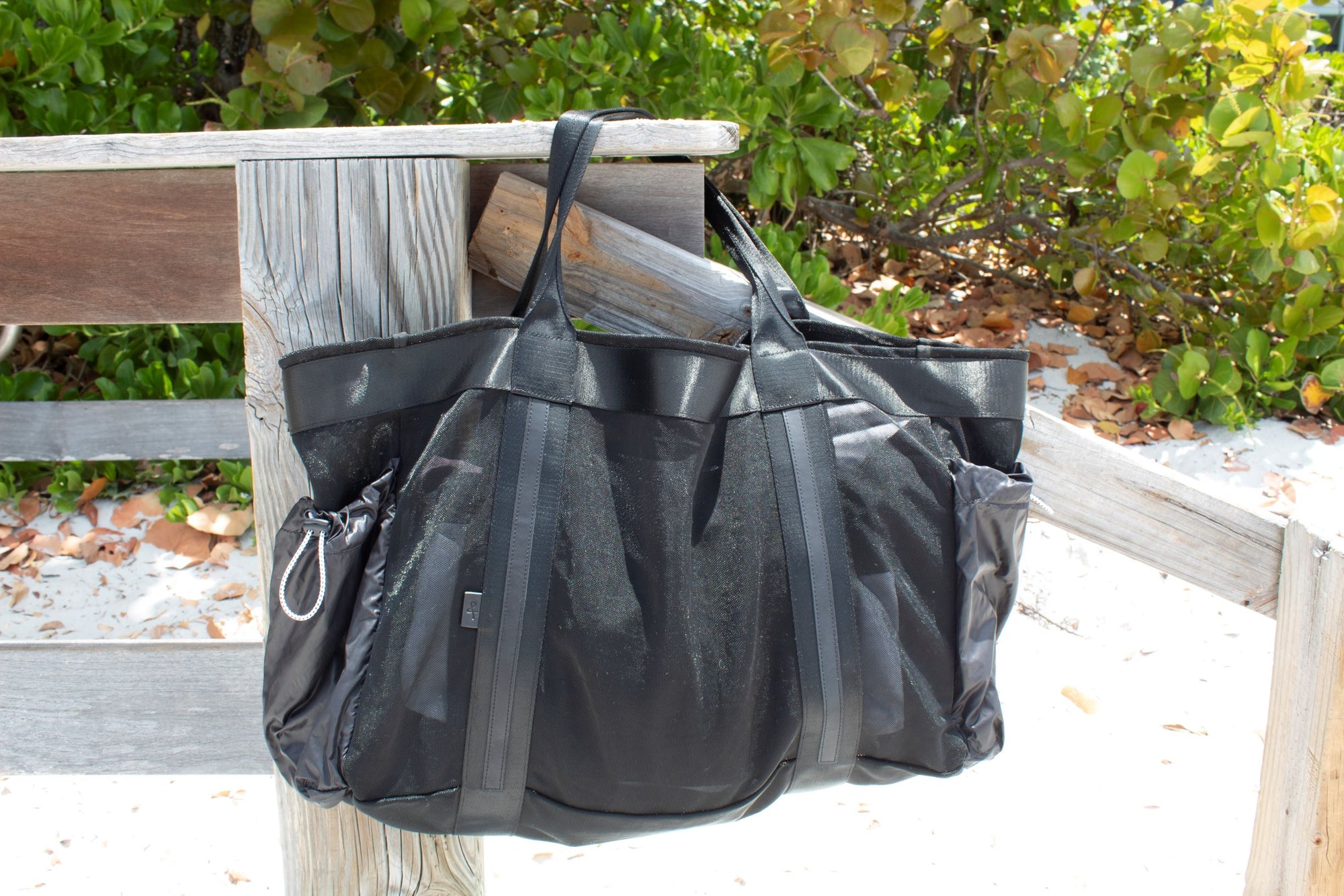 Large black mesh beach bag with nylon side pockets hanging off dock post.
