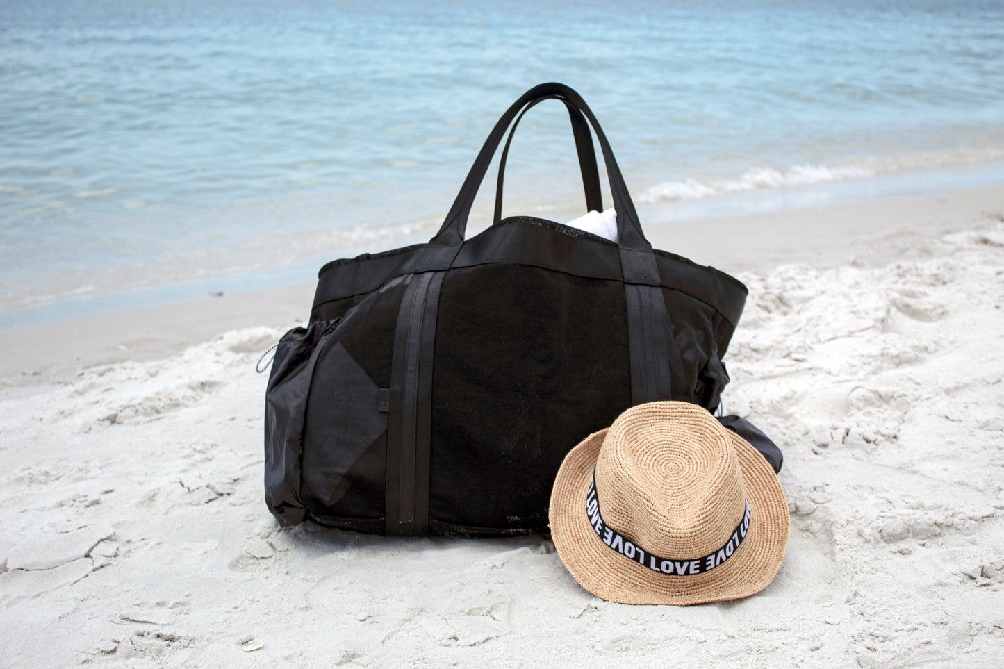 Large black mesh beach bag with nylon side pockets and straw Essential Hat with LOVE band sitting on beach.