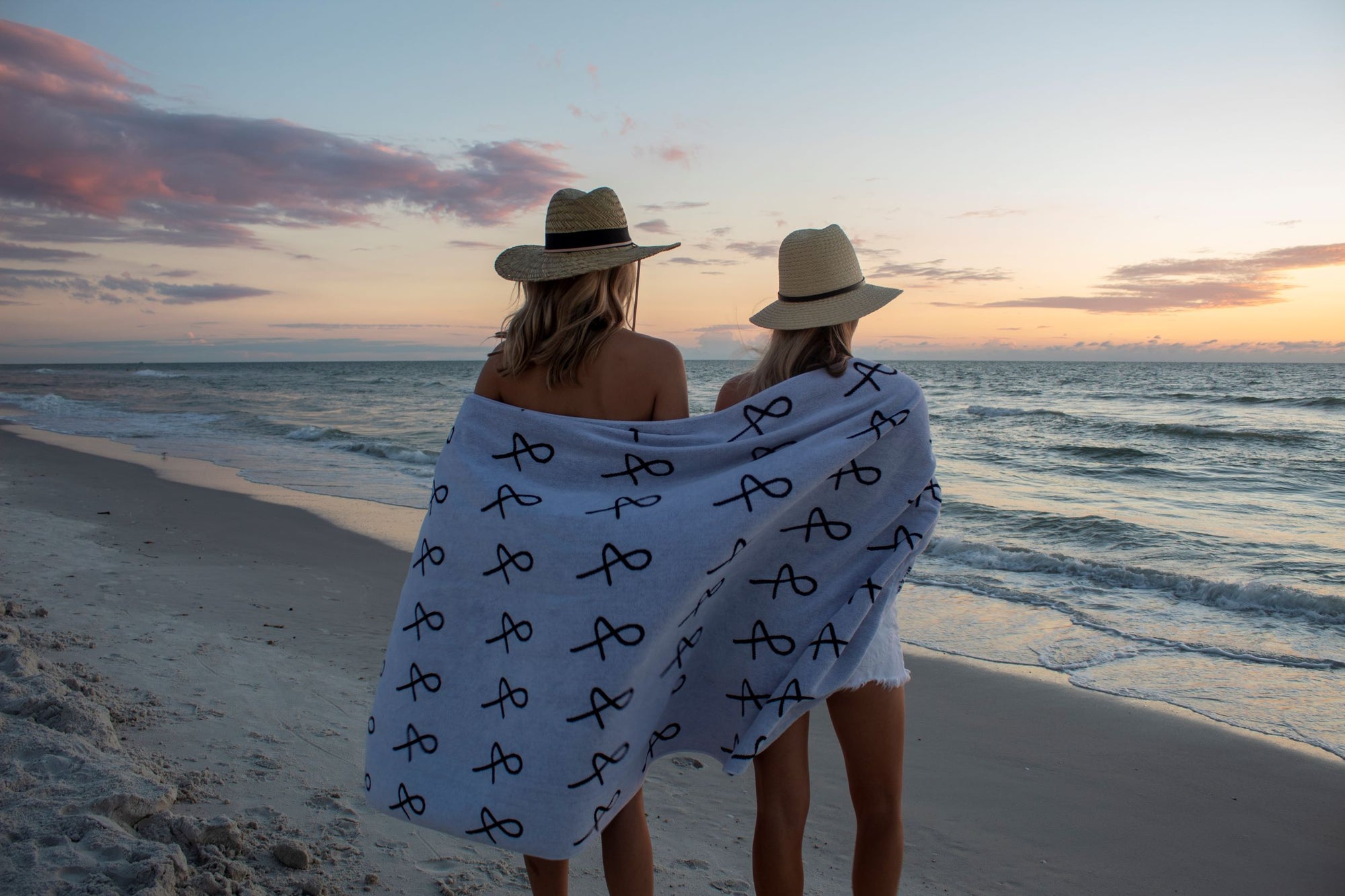 Two people at beach with large Anya & Niki beach towel wrapped around them, wearing straw hats.