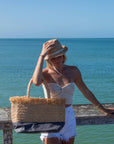 Person at beach holding natural raffia straw tote with linen lining and gray color block base and wearing a raffia straw hat with rope band.