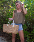 Person holding the Anya & Niki Augustine straw bag - a midsized hyacinth straw tote with cream leather handles and linen cinch top.