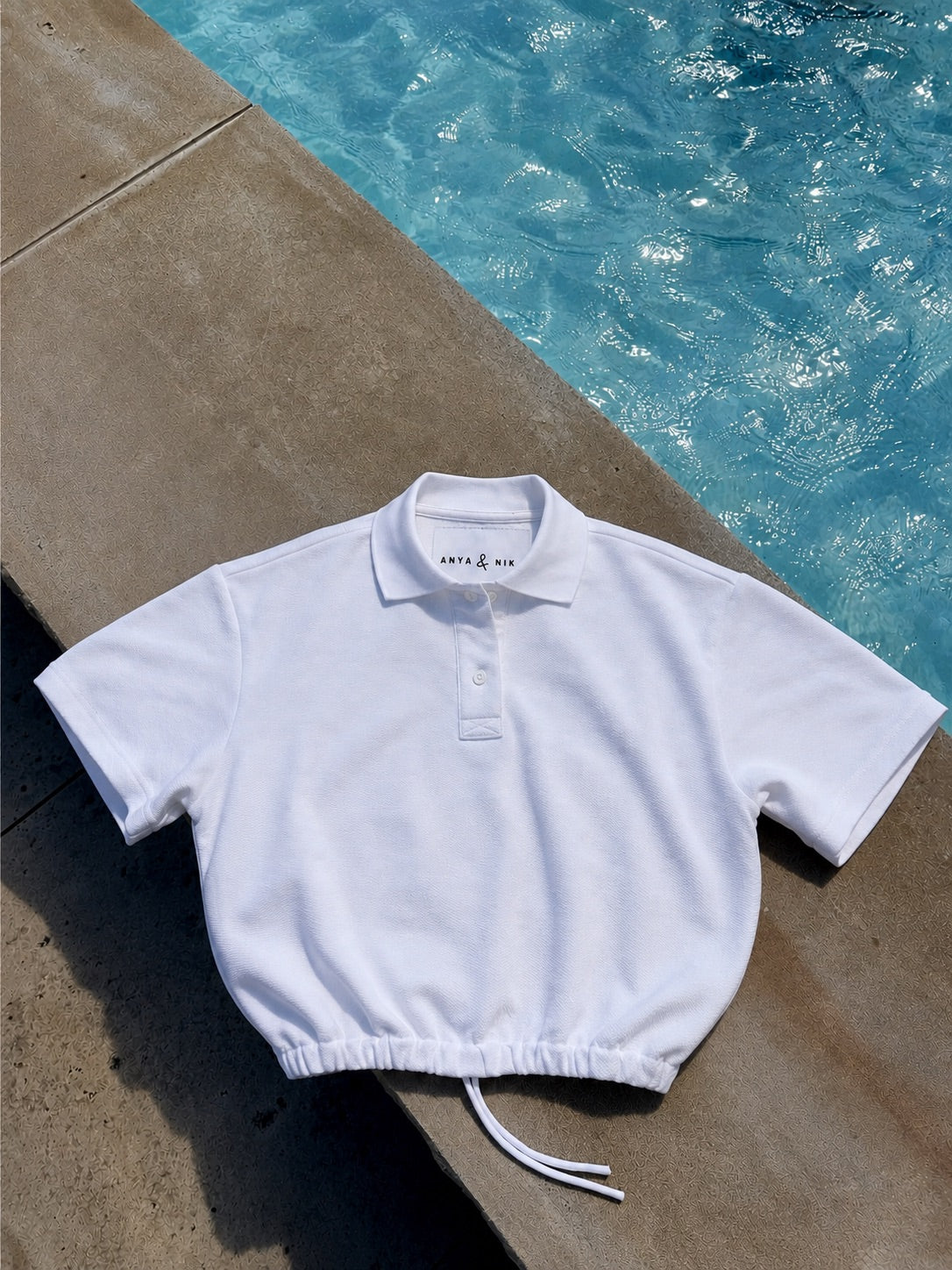 A white short-sleeve polo shirt with a drawstring hem, laid flat on a concrete poolside surface with blue water in the background.
