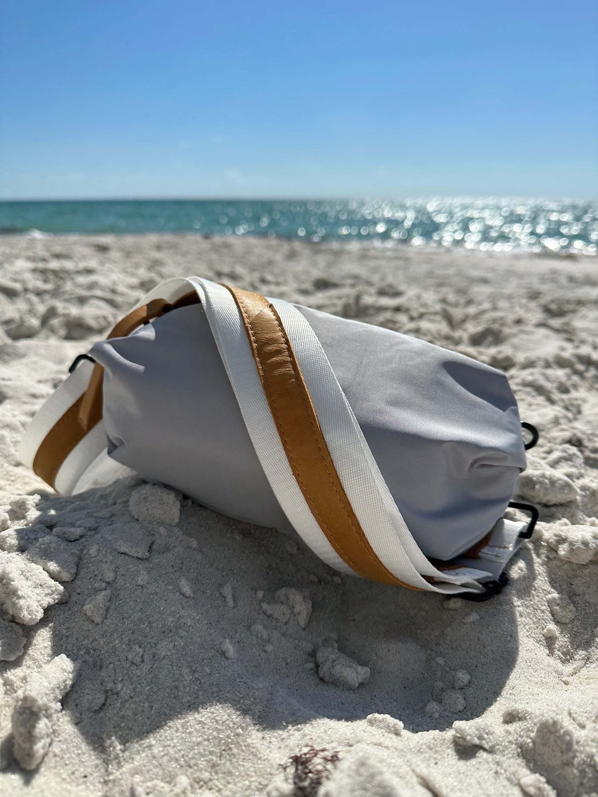 Gray nylon belt bag with white crossbody strap and tan leather detail on the beach