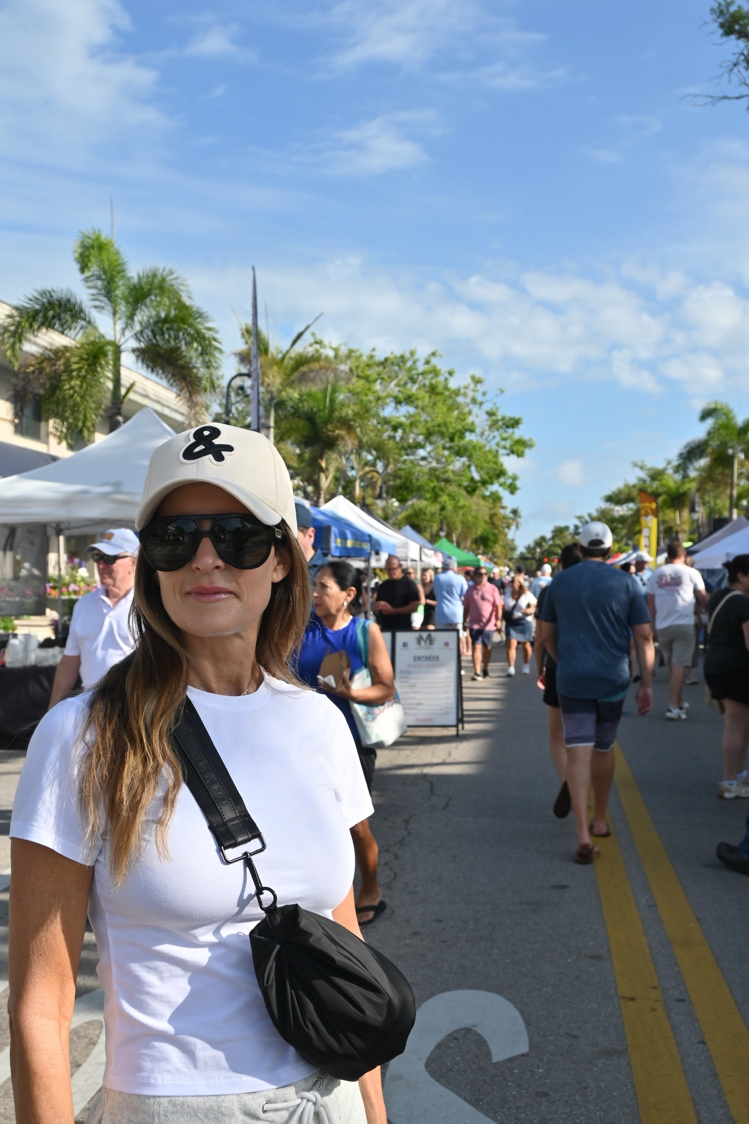 Woman wearing a white cap with a black logo and sunglasses, standing on a street with palm trees and people in the background.