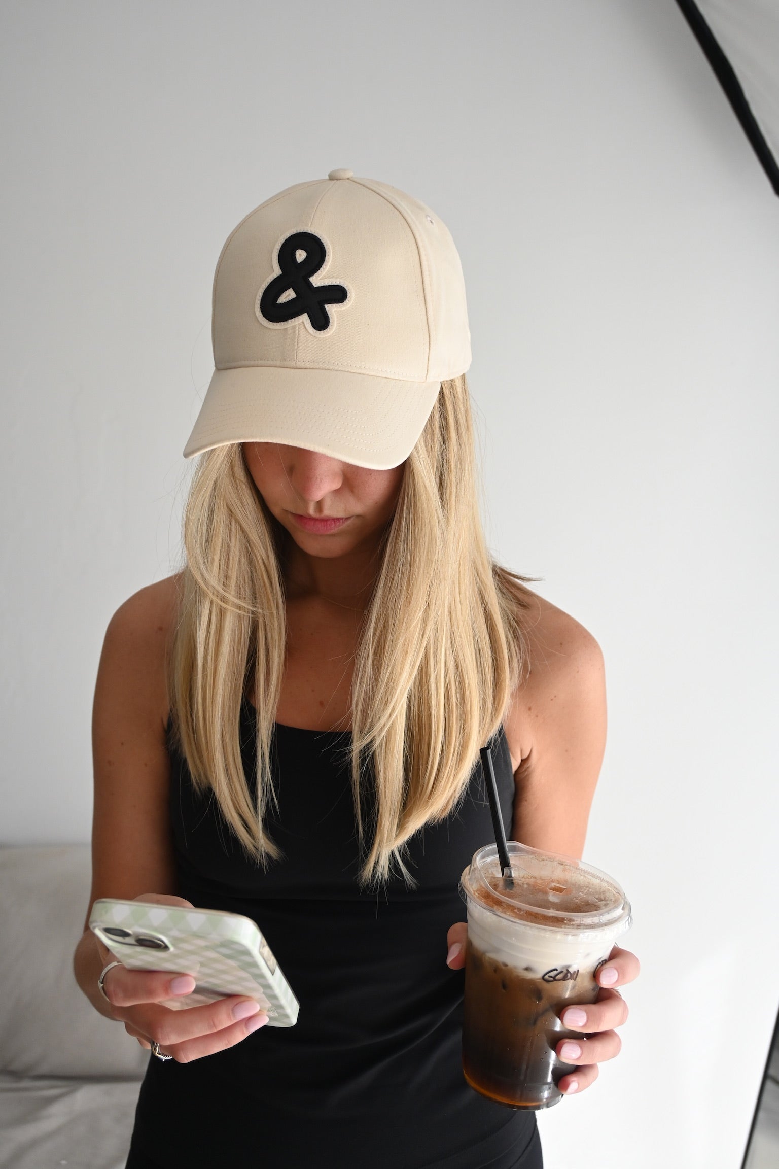 Person wearing a beige cap with an ampersand symbol, holding a phone and a cup of iced coffee.