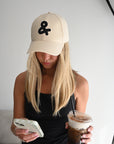 Person wearing a beige cap with an ampersand symbol, holding a phone and a cup of iced coffee.