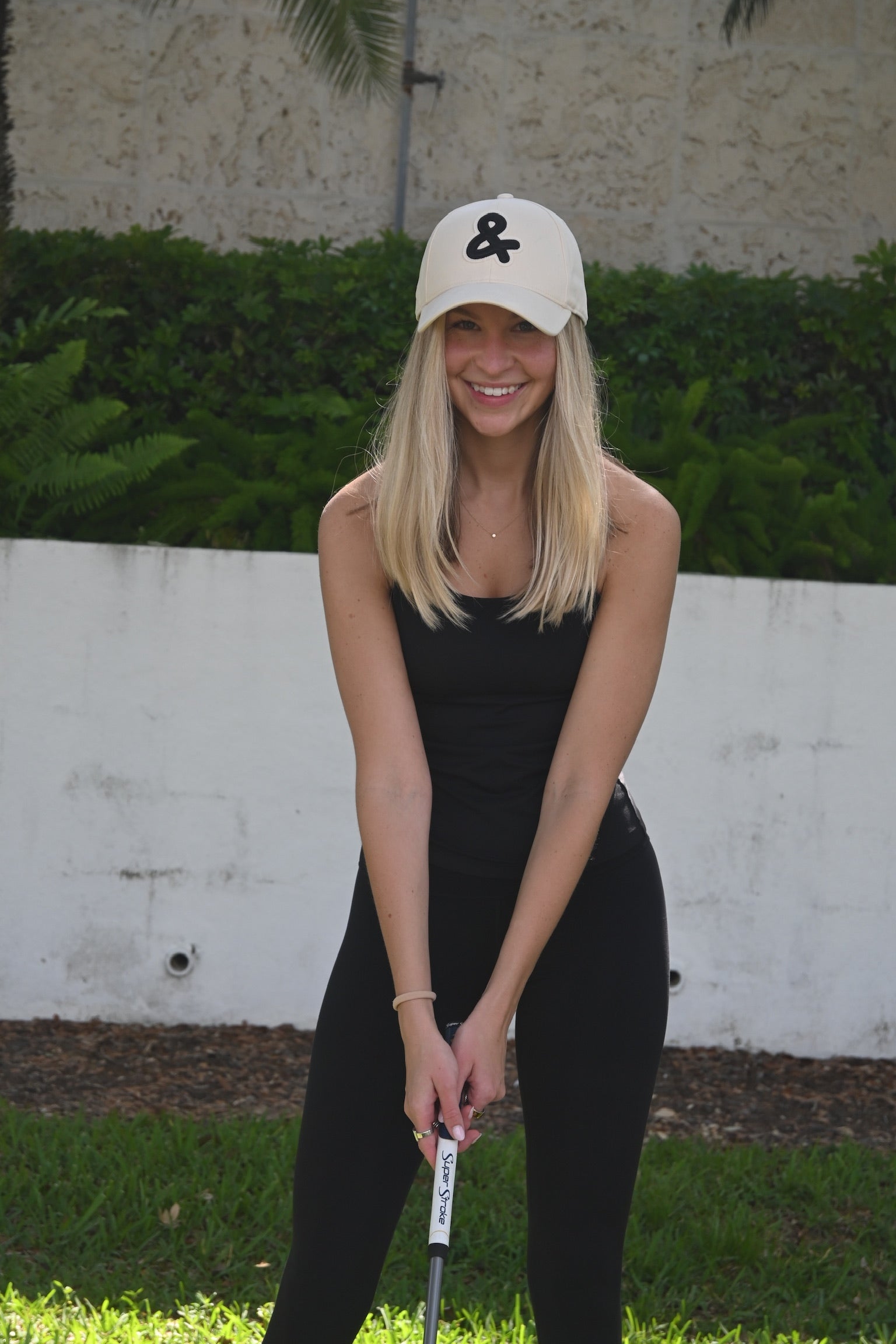 Woman in black outfit and white cap holding a golf club on a grassy area with a wall and plants in the background.
