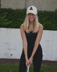 Woman in black outfit and white cap holding a golf club on a grassy area with a wall and plants in the background.
