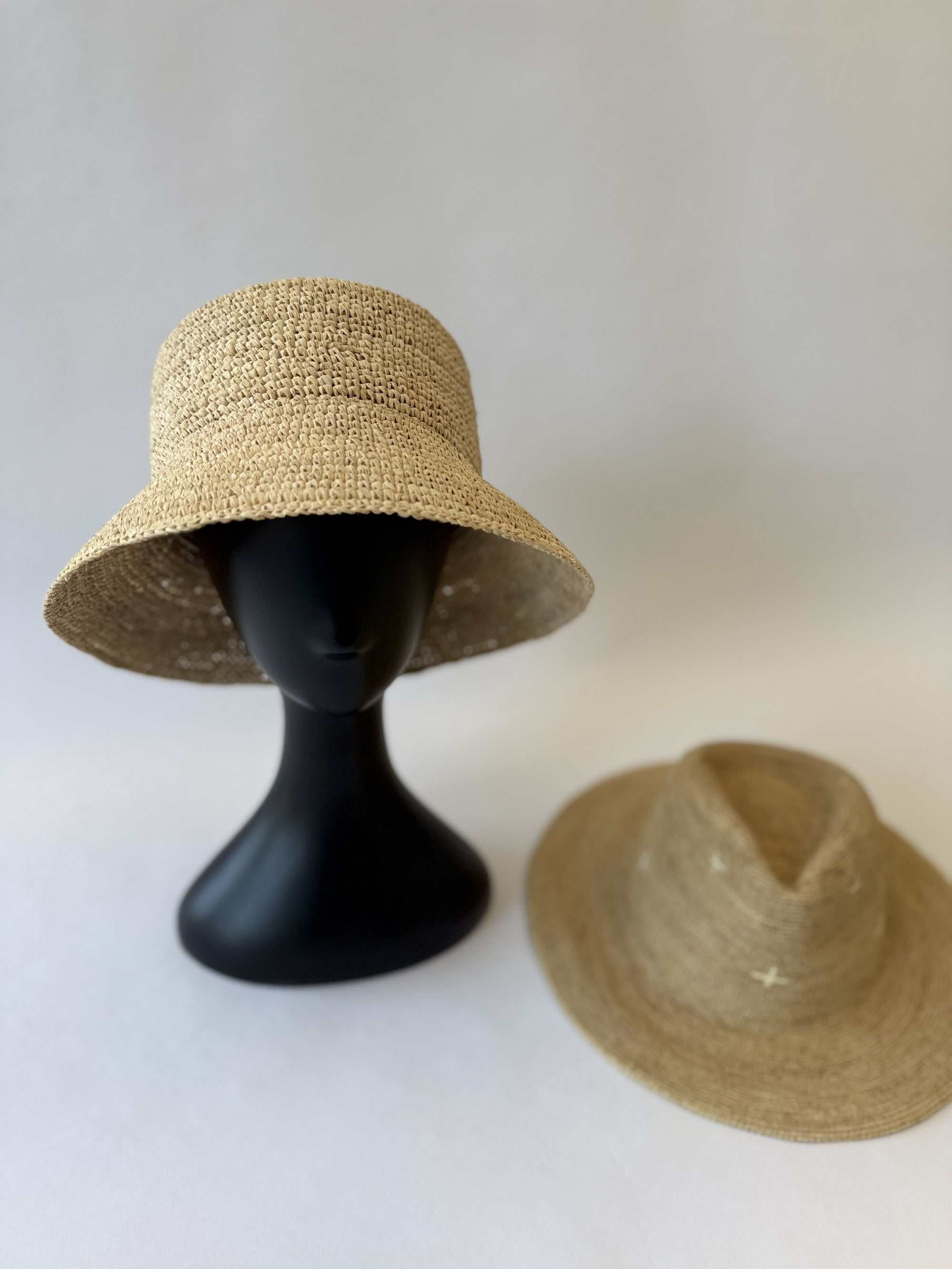 Two straw hats on a white background, one on a black mannequin head and the other on a flat surface.