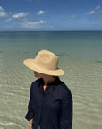 Person wearing a straw hat and dark shirt standing on a beach with clear water and blue sky.