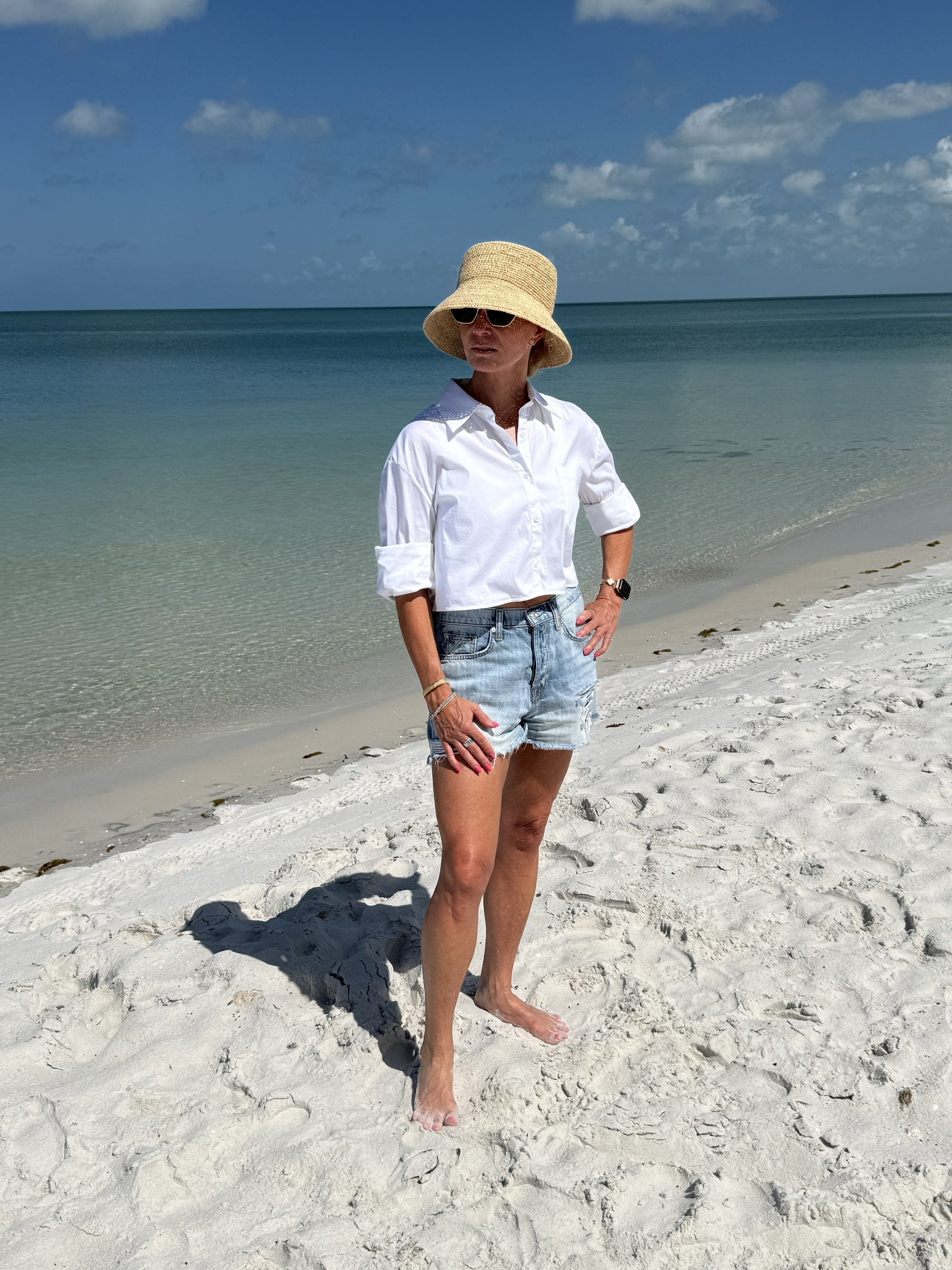 Person wearing a straw bucket hat standing on a sandy beach with ocean and blue sky in the background