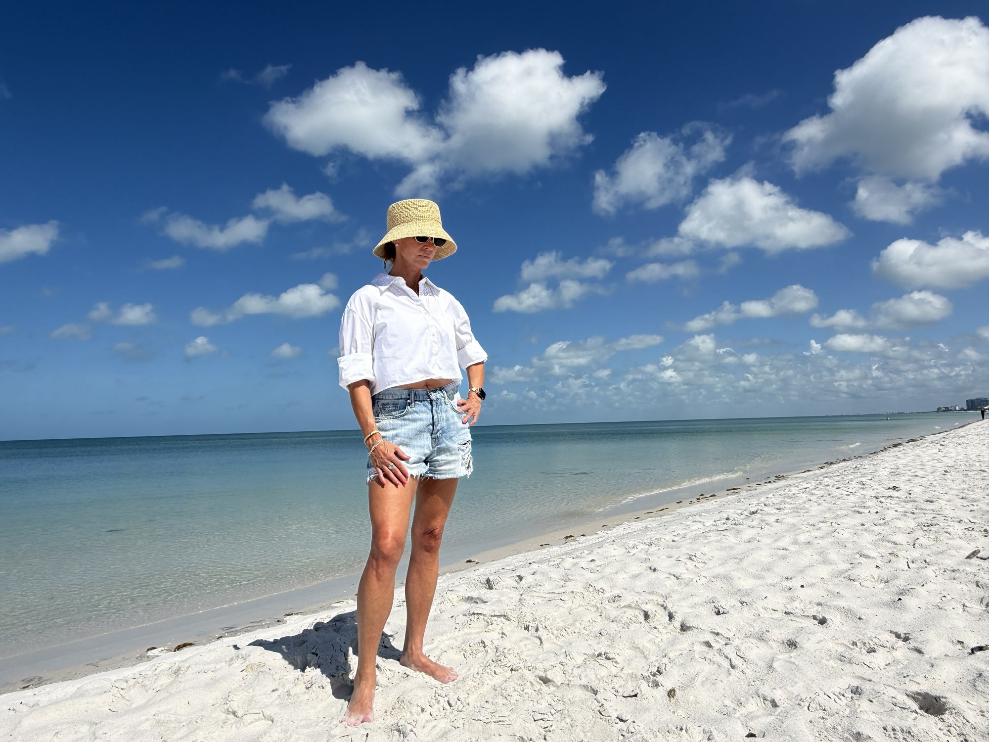 Person wearing a straw bucket hat standing on a sandy beach with ocean and blue sky in the background