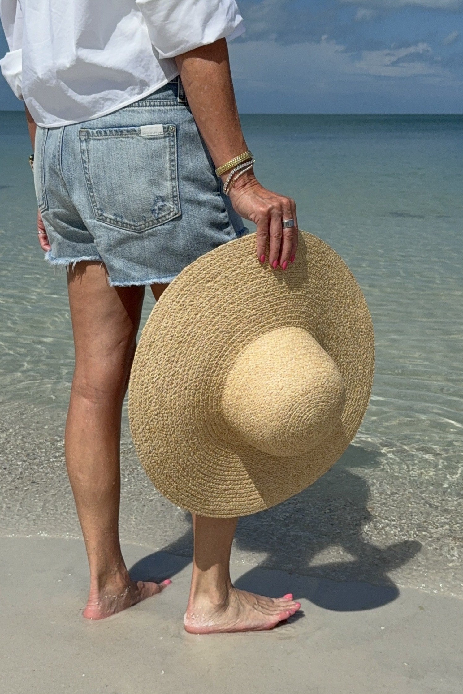 Person holding a straw hat on a beach with clear water and blue sky.