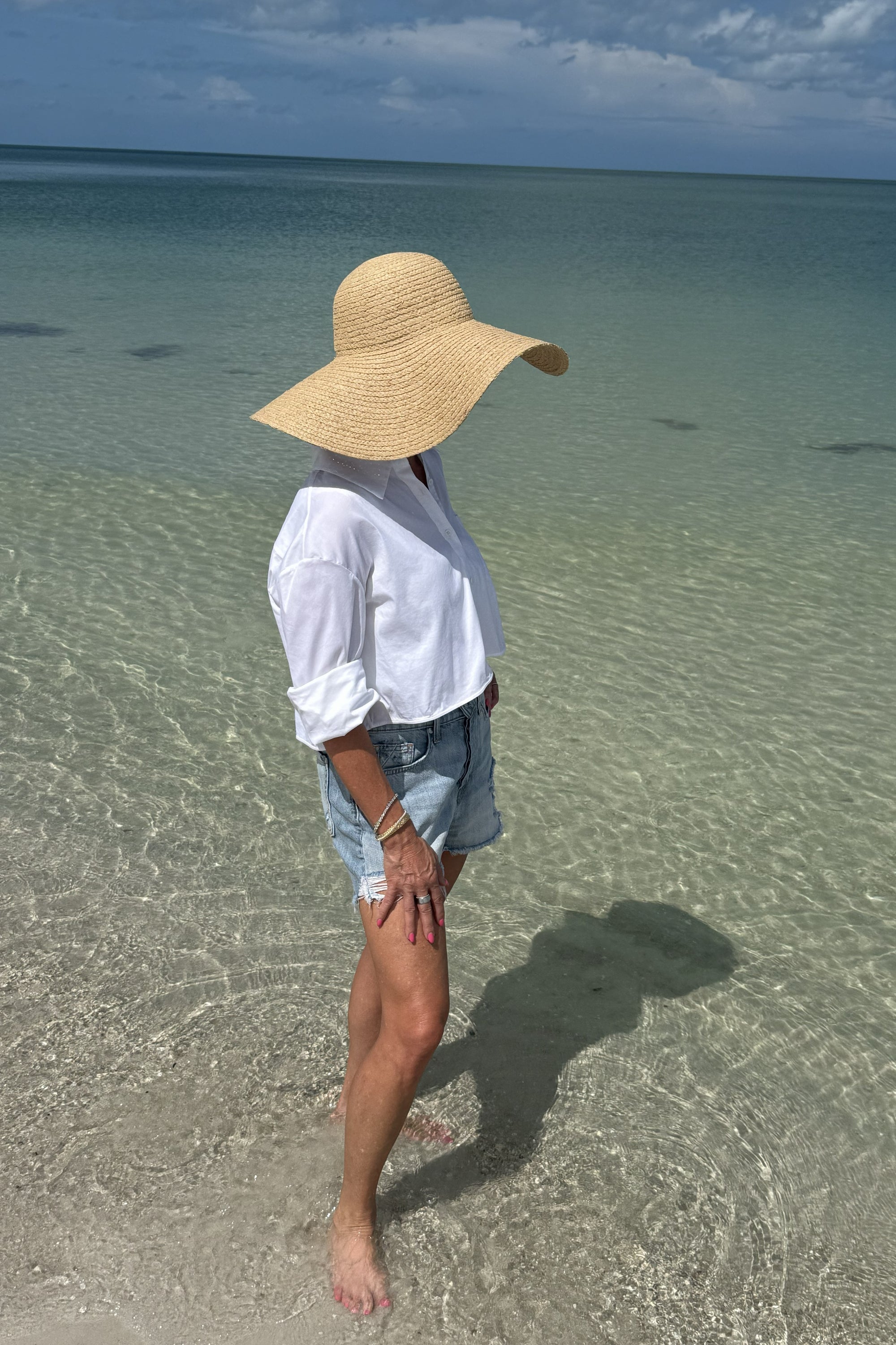 Person wearing a wide-brimmed hat standing in shallow water on a beach.