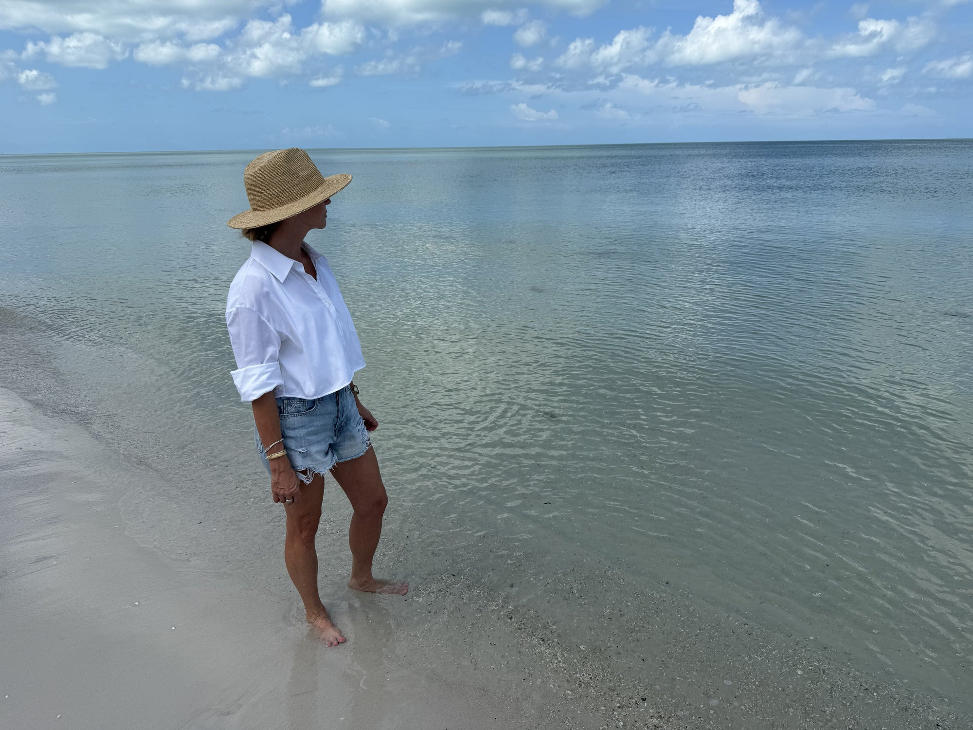 Person standing on a beach wearing a straw hat with clear water and blue sky