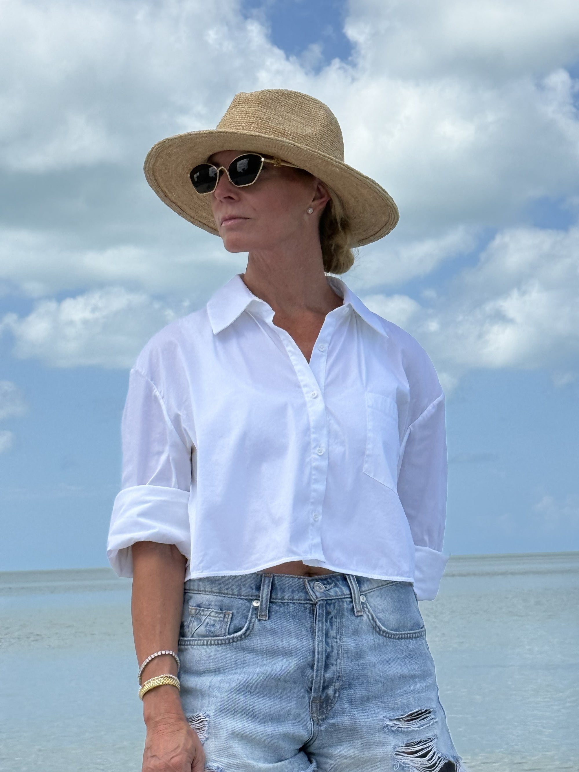 Person wearing a white shirt, straw hat, and sunglasses on a beach with blue sky and ocean.