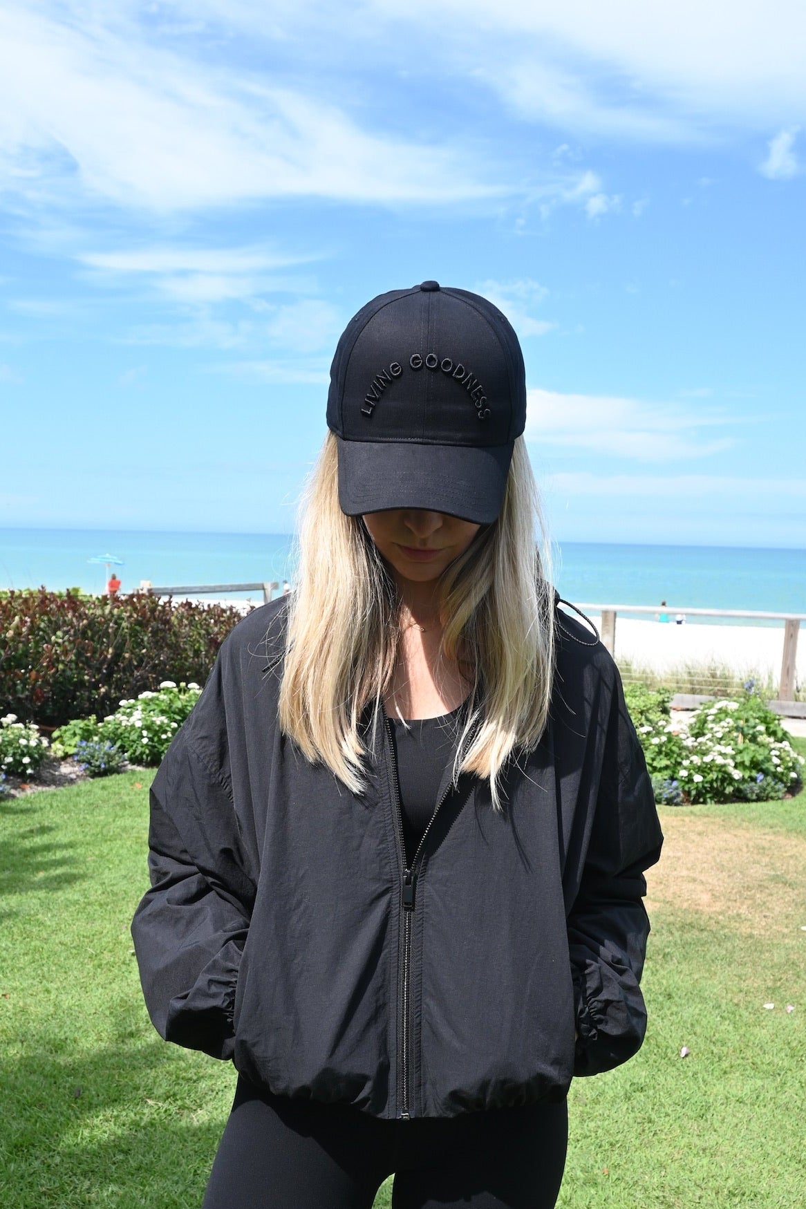 Person wearing a black jacket and black baseball cap standing on grass with ocean in the background